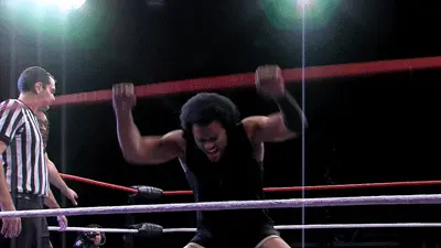 A wrestler in a black outfit, known as the Gambler, pounds the mat with both fists inside a wrestling ring as a referee stands nearby; bright overhead lights shine in the background.