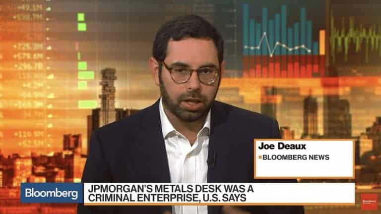 A man with dark hair, beard, and glasses appears on a news broadcast. Seated against a backdrop of financial graphs and stock market data from JP Morgan, his image is partially obscured by a grey bar at the bottom.