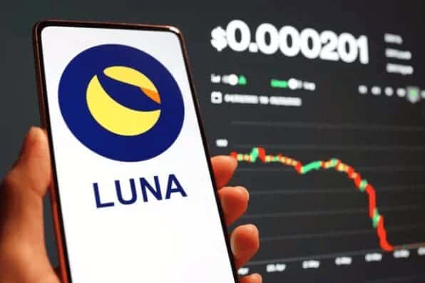 Uma pessoa segura um smartphone exibindo o logotipo da criptomoeda Terra LUNA. No fundo, uma tela mostra um gráfico de linha em declínio e o número 0,000201, sugerindo uma queda no valor. 