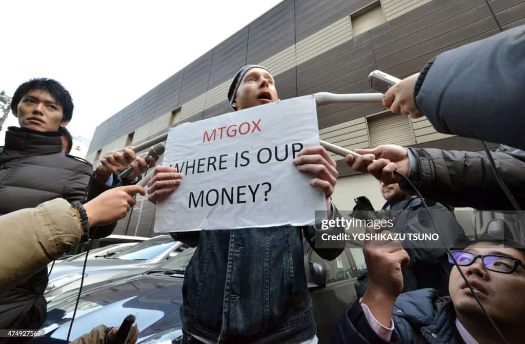 A person holds a sign reading "MTGOX WHERE IS OUR MONEY?" while being surrounded by several people extending their hands with recording devices and microphones, highlighting the Mt. Gox fud. It appears to be an outdoor setting near a building. Image credit: YOSHIKAZU TSUNO.