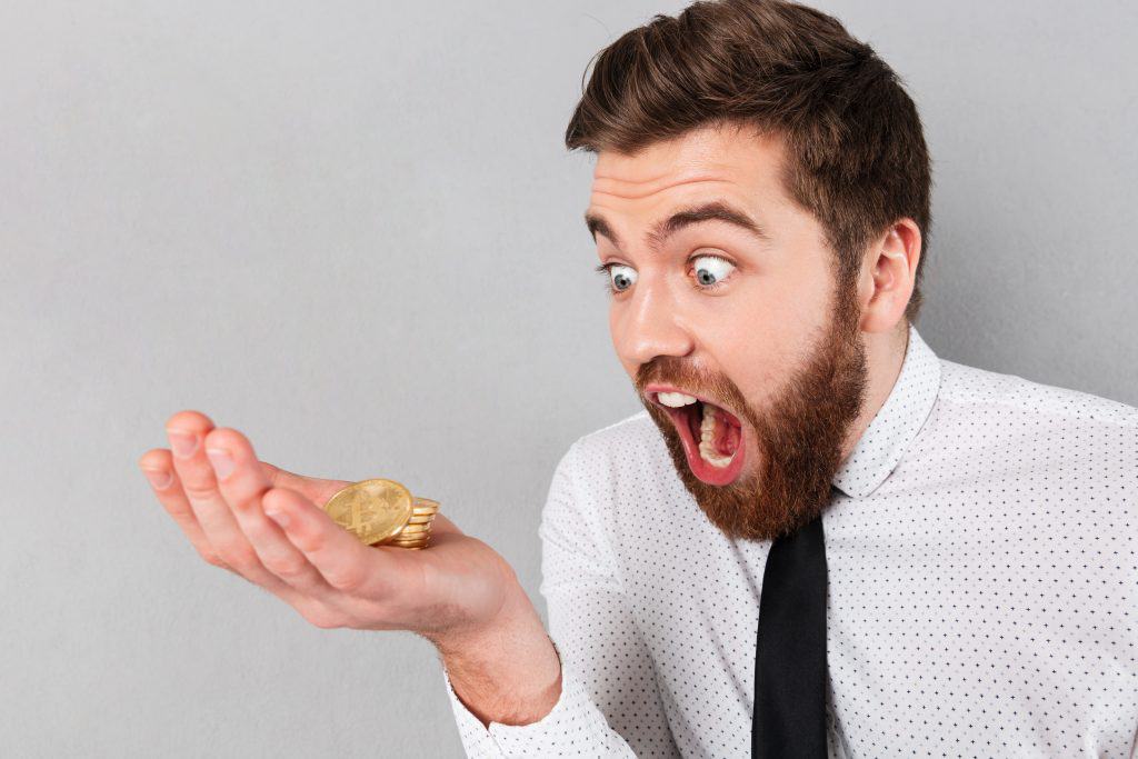 A bearded man in a white shirt and black tie looks surprised while holding several gold coins, perhaps realizing his recent crypto trading mistakes. The background is a plain gray wall.