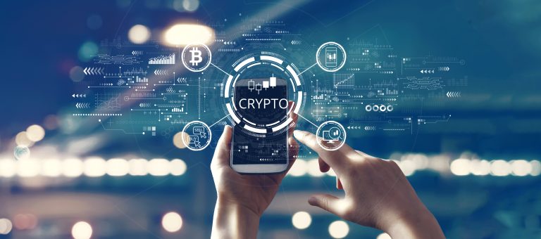 A person holds a smartphone displaying the word "CRYPTO" surrounded by digital cryptocurrency icons and symbols against a blurred cityscape background.