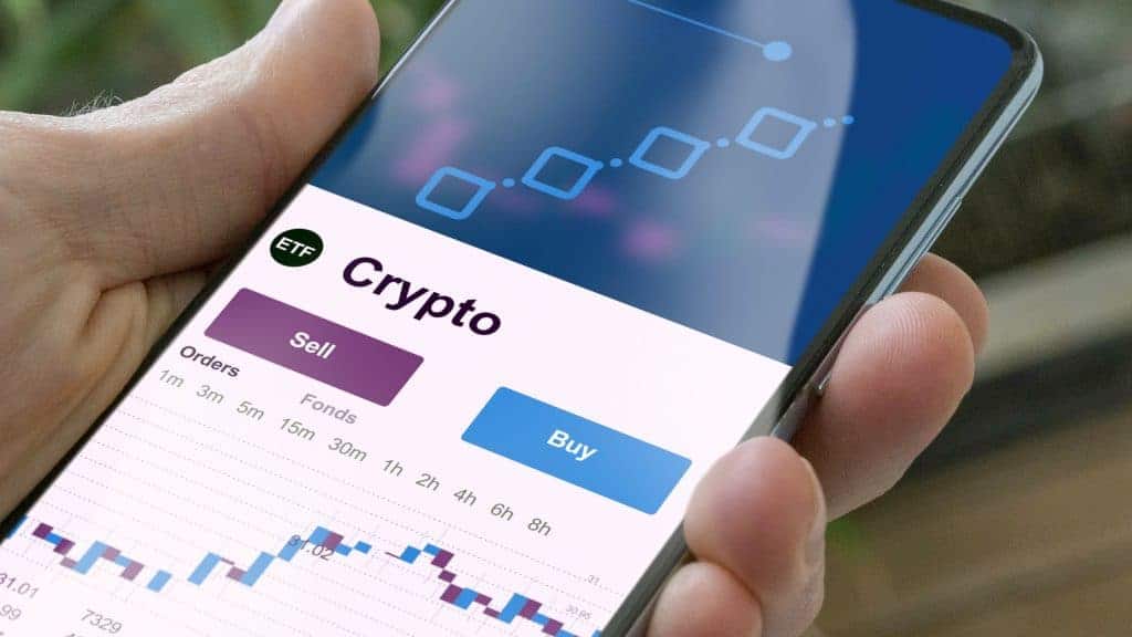 Uma pessoa segura um smartphone exibindo um aplicativo de trading de criptomoedas, com as opções “Crypto”, “Vender” e “Comprar”, um gráfico e intervalos de tempo visíveis na tela variando de 1h a 8h.